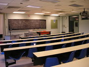 view of a classroom from the rear, with blackboard and three desks and tables at the front of the class, and five rows of long curved student desks with blue chairs attached.