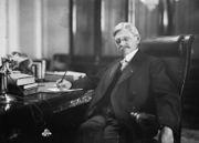 A man sitting at a desk looking directly at the photographer; one hand is holding a pen to a document on his desk while the other is hanging over the arm of his high backed chair, his legged streched forwards, he is wearing a suit, neck tie, and glasses, and has a serious expression.