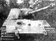 A head-on view of a large tank with a flat faced turret. Its sloped bow armour is scarred with several fist-sized dents, and there is a fist-sized hole in the front of the turret