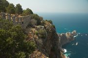 Blue-green sea surrounds a rocky peninsula covered by green trees and a stone castle wall with crenelations.