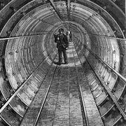 Etching of a circular tunnel made of bolted segments. A walkway of boards runs down the centre of the tunnel and a man in workman's clothes with a bag over his shoulders stands in the middle distance