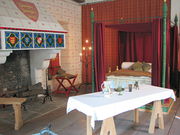 On the left is a fireplace with various heraldic arms painted on it, on the right is a four-post bed, and in the front is a set table on trestles. The floor is wooden and the walls are covered with painted patterns and drapes.