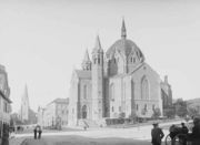 Large, fanciful building with towers, pointed arches, and a large dome in the in the center, topped by a spire.