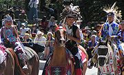 "Colour photograph of Tsuu T'ina children in traditional costume on horseback at a Stampede Parade in front of an audience"