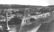 A grainy, black and white photograph showing several single story buildings next to a dirt road.  Several homes and other buildings stretch toward the mountains in the distance.  A chimney is seen in the foreground.