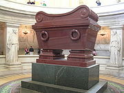 Photo of a large, shiny burgundy cuboid-shaped vessel raised on a dark green plinth. There are two female statues in the background either side of the vessel.