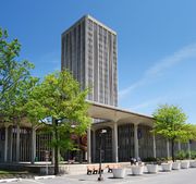 A single modern-style tower is surrounded by a lower open-air pavilion with trees accenting the area.