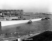 A large hull being pushed through a shipyard by tugboats.