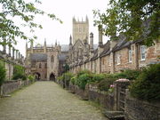 Street scene. Terraces of old red brick houses on either side of the road. At the far end is a building with arches and small tower. Beyond can be seen the cathedral tower.