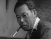 Photo of Takashi Shimura, a middle-aged man with short hair, facing left and staring pensively at the ground; he wears a suit jacket and holds a cigarette in a cigarette holder in his mouth.