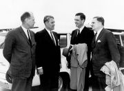 Four men in suits are outdoors, speaking to each other in front of a large white automobile.