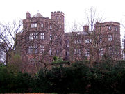 Seen from a low angle through the branches of trees is a substantial stone house with two-three storeys, bay windows and some castellation.