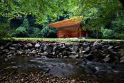 A small, boxy, wooden stage with a trapezoidal overhang stands in the center of meadow. In the foreground is a running stream with a stone embankment.