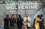 Atop a stage are three man in black clothing, Liev Schreiber wearing a gray jacket and black pants, and Lynn Collins, wearing a yellow dress, hugging will.i.am, who is in black clothing. In the background is a billboard reading "X-Men Origins Wolverine: World Premiere - Tempe, Arizona. Colored paper flies through the stage.