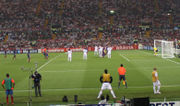 A man in a blue-and-red shirt kicks a football past a row of men in white shirts towards a goal.