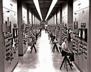 A long hallway, with bulky computers reaching from the floor to the ceiling, covered in switches and dials. Women sit on chairs operating the machinery.