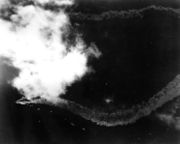 A distant overhead view of a patch of ocean; towards the left, partially obscured by a patch of cloud, is a large warship. The ship's wake trails out in a zig-zag pattern behind her.