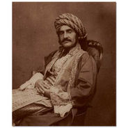 Sepia photograph of a man in 19th century Middle Eastern dress, with a large moustache, reclining in a chair with his hands crossed across his lap.