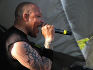 A man, with a large tattoo on his right arm, sings into a microphone while looking at the audience.