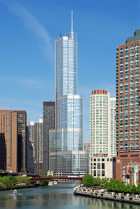 Distant ground-level view of a 100-story skyscraper with a blue, all-glass facade; the building has setbacks ant many levels, and a thin spire rises above its roofline.