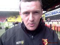 The head and shoulders of a man in his thirties. He has short hair and is wearing a black tracksuit top. A grass field and two sides of a sports stadium are visible in the background.