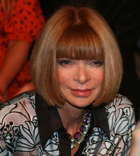 A woman with brown bobbed hair and green eyes wearing a print top with black collar and two necklaces with blue and purple stones