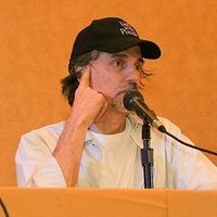 Upper body shot of man sitting behind desk with baseball cap speaking into a microphone