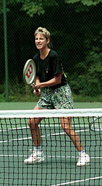 A blonde-haired female tennis player with multi-colored shorts and a black shirt, with the tennis racket out in front of her