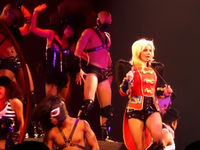 Image of a blond woman. She is standing with a red feathered jacket, carrying a whip around her neck and singing in a wireless microphone. Several people surround her, all wearing S&M outfits.