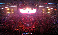 Faraway image of an arena full of people. In the middle, there is a stage composed by three rings, a big one in the middle and two smaller rings on the sides. A 360 degrees cylinder shaped screen is hanging from the ceiling along with different subwoofers and speakers.