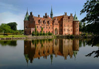 A large three-storied castle has  steeply pitched roofs,  two circular towers and a spire, and a large arched entrance facing a lake which reflects the castle's image.