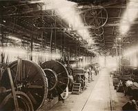 A photograph showing the interior of a large factory building with lighting provided by overhead skylights, underneath which is suspended a line shaft providing power via pulley belts to various machines on the factory floor, some of which dwarf workers standing by their stations