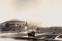 A photograph showing workers spreading or gathering coffee beans drying on a large paved plaza with an elegant, two story neoclassical building on the left and warehouses and other plantation buildings in the background