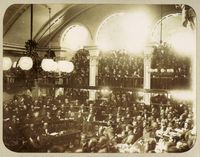 A photograph showing a large number of men seated on semi-circular tiers in a vaulted chamber as a large crowd looks on from an arcaded balcony