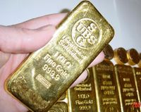 A hand holding a gold bar surrounded by several gold bars and other gold pieces in the background.