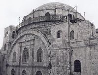 A large domed roof sits upon a bricked parapet, which includes arched windows, three of which are visible. The base of the parapet is surrounded by a narrow veranda which is fenced with decorative metal grating. Below this, a set of three large arched windows, on two levels, are situated under a large stone arch, itself flanked by two supporting towers. The left tower consisting of an upper section is slightly taller that its counterpart on the right.