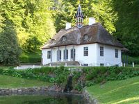 A thatched and plaster country house sits on a rise with trees in the background. The roof has three gables and a columned cupola with a point top. Steps go up to the three arched paned glass entrances on the columned porch.