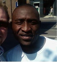 A black man with a shaved head smiling directly at the camera. He is wearing a white t-shirt and is standing on a street next to a fair haired man who is partially visible.