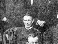  A man, fresh-faced with dark, brushed-back hair, seated among a group. He is wearing a naval officer's uniform with a high, stiff collar