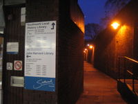 Evening scene looking into an alleyway, bounded on the right by a long, 15-foot-high brick wall with brightly lit lamps hanging on it. On the left, there is a building with a sign saying "John Harvard Library", with telephone numbers and opening hours. An arrow points toward the entrance to the library in the alleyway.