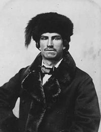 Black and white photograph of a man with a short moustache and earings, wearing a fur lined dress jacket, bow tie and fur hat