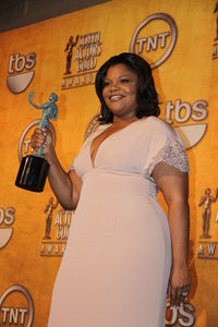 An African American female with dark brown hair that reaches her shoulders. She is wearing a short sleeved light pink dress and a ring on her left hand. She is holding a greenish-blue statue that has a bronze plank with her right hand. In the background, there is an orange wall with logos and writing, such as the words "tbs" and "TNT" (which has a circle around it).