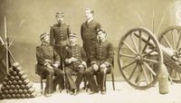 A photograph depicting a group of 5 uniformed men posed between a pyramid of artillery shells on the left and a wheeled field artillery piece on the right