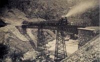 A photograph showing a steam locomotive pulling freight cars, passing across an iron tressle which spans a gorge with a rushing stream at its bottom