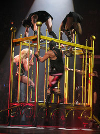 The right profile of a female blond performer. She is moving inside a giant golden cage. She is wearing a black corset with diamonds and fishnet stockings. Four leather-clad men seem to be chasing her.