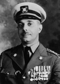 Head and shoulders of a man with a small moustache wearing a military uniform with many medals on his chest. He is looking into the camera, smiling slightly.