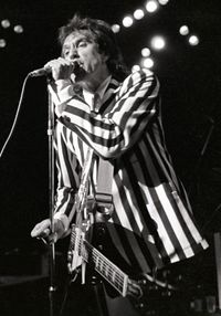 A man stands onstage with a guitar strapped across his chest. It hangs limp and unused, as he is focusing on singing into a microphone directly in front of him, which he grasps with his left hand. He wears a black-and-white, vertically striped suit.