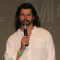 A man talks to an audience using a microphone held in his hand. He has a light complexion, full beard and long brown hair.