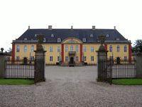 An iron gate opens on to a two-story rectangle palace with large, attached wings. Rounded arch windows and a stone work over the entrance and on the second floor of the wings.