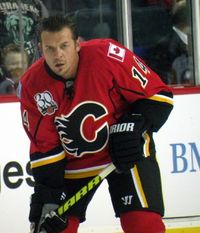 An ice hockey player stands partially crouched, leaning on his stick.  He has short black hair and is not wearing a helmet.  He is wearing a red uniform with a large black C on his chest.